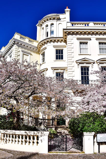 A white, multi-story Victorian-style building with ornate architectural details, including bay windows and decorative cornices, set against a vivid blue sky. In the foreground, blooming cherry blossom trees with pink flowers partially obscure the building, and a stone staircase leads up to the entrance, which is gated. The image captures a bright, sunny day, highlighting the clean and well-maintained exterior of the property, characteristic of the residential area around Pembridge Villas, Notting Hill, W10. Carpet Cleaners W10 specializes in surface cleaning and deep cleaning services for such elegant homes, ensuring hygiene and maintenance are kept to a high standard.