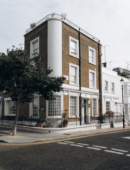 Exterior view of a multi-story residential building in Pembridge Villas, Notting Hill, W10, showing a brick façade with white window frames and decorative trim. The building features a corner design with a rounded white wall section, a rooftop terrace with a metal railing, and potted plants near the entrance. The surrounding sidewalk is lined with a black metal fence, and a nearby tree provides some greenery. The scene captures the clean, well-maintained appearance of the property on a cloudy day, reflecting a typical residential setting in W10, with no interior surfaces or cleaning tools visible. For professional surface cleaning and deep cleaning services tailored for residential properties in Pembridge Villas, consider contacting Carpet Cleaners W10.