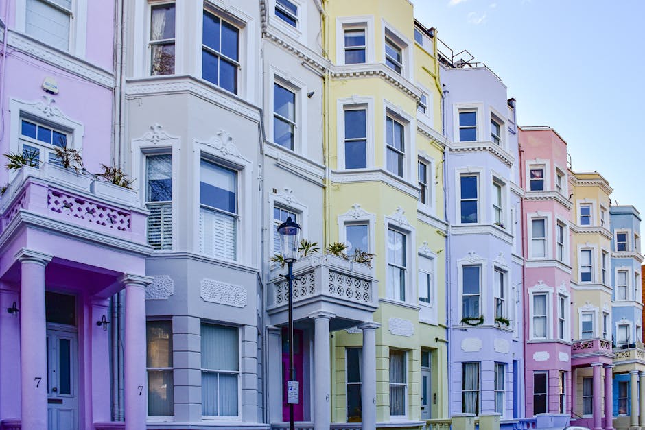 A row of pastel-colored residential buildings with multiple floors, featuring large rectangular windows, decorative plaster moldings, and small balconies with wrought iron railings. The buildings exhibit a mixture of yellow, pink, lavender, and white facades, with some windows adorned with flower boxes. The ground level showcases entrance doors and pillars, with a street lamp positioned in front. The scene is illuminated by natural daylight, emphasizing the clean and well-maintained appearance of the facades, aligning with professional surface cleaning and maintenance practices typical of domestic property care. Carpet Cleaners W10 specializes in thorough cleaning and sanitisation of residential surfaces, ensuring hygienic living environments in properties like these.