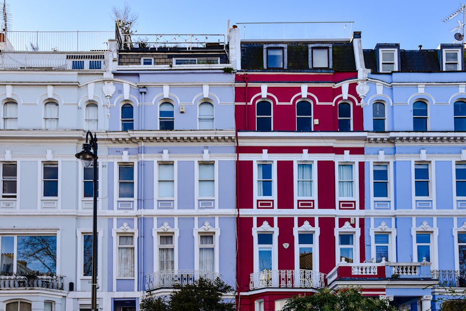 Photograph of a row of Victorian-style terraced houses in Notting Hill, featuring colorful facades with detailed white moldings and decorative elements. The buildings are painted in pastel blue and vibrant red, with multiple large windows fitted with white frames, some with curtains visible inside. The rooftops are visible at the top, with small dormer windows and a rooftop terrace with a railing. A black street lamp stands in front of the buildings. The scene is well-lit with natural daylight, emphasizing the clean and well-maintained appearance of the exterior surfaces, reflecting a focus on preservation and aesthetic appeal relevant to surface cleaning and maintenance. This image visually aligns with the theme of property care, which is rooted in effective cleaning and sanitisation, as discussed on the page about permits for building cleaning in Notting Hill, W10, by Carpet Cleaners W10.