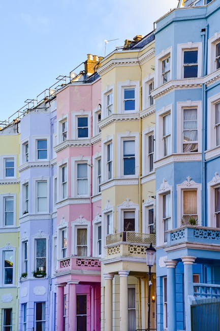 A row of pastel-colored residential buildings with multiple floors, featuring large rectangular windows, decorative plaster moldings, and small balconies with wrought iron railings. The buildings exhibit a mixture of yellow, pink, lavender, and white facades, with some windows adorned with flower boxes. The ground level showcases entrance doors and pillars, with a street lamp positioned in front. The scene is illuminated by natural daylight, emphasizing the clean and well-maintained appearance of the facades, aligning with professional surface cleaning and maintenance practices typical of domestic property care. Carpet Cleaners W10 specializes in thorough cleaning and sanitisation of residential surfaces, ensuring hygienic living environments in properties like these.