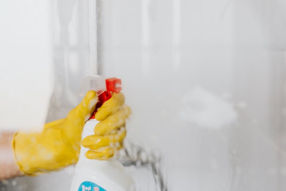 A close-up view of a person's hand wearing a yellow rubber cleaning glove, holding a white spray bottle with a red nozzle, actively spraying cleaning solution onto a glass surface, likely a kitchen or bathroom shower door or window, which appears slightly fogged or smudged. The background features a white tiled wall and a partially visible mirror or reflective surface, illuminated by soft, natural light, signaling a domestic setting focused on surface cleaning and sanitisation for effective hygiene maintenance. Carpet Cleaners W10 specializes in professional domestic cleaning and deep cleaning services in the Notting Hill area, including emergency stain removal and surface sanitisation.
