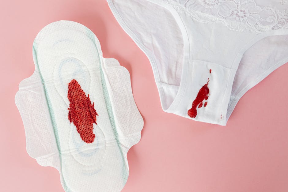 A menstrual pad and a pair of white cotton underwear with visible bloodstains on a soft pink background, illustrating a typical scenario requiring urgent surface cleaning and sanitisation. The pad is laid flat, showing the blood stain at its center, while the underwear is partially folded with a noticeable blood mark near the waistband. The scene highlights the importance of professional deep cleaning and hygiene maintenance for domestic and commercial environments, as promoted by Carpet Cleaners W10. The soft lighting enhances the clarity of the stains and the fabric textures, reflecting a focus on effective stain removal and surface sanitisation in line with emergency stain removal services in Notting Hill, W10.