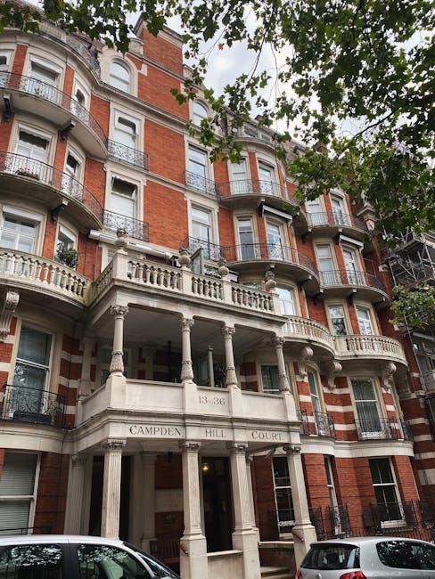 Exterior view of a multi-storey residential building on Campden Hill, Notting Hill, with a combination of red brick walls and white stone detailing. The building features ornate balconies with decorative iron railings, large sash windows, and a grand entrance supported by classical columns. The area is surrounded by lush green leafy trees, with partial views of parked cars at the street level. The lighting appears natural, highlighting the building's architectural details and the well-maintained façade, reflecting a typical setting for a property that may require interior and surface cleaning as part of estate maintenance in accordance with the estate cleaning checklist for Royal Crescent, Notting Hill, W10. Carpet Cleaners W10 specializes in deep cleaning and sanitisation services to uphold hygiene standards for residential properties.