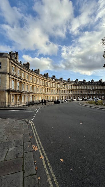 An exterior view of a curved, historic sandstone building with multiple columns and ornate architectural details, situated along a paved street in an affluent residential area. The scene features a partly cloudy sky with patches of blue, casting soft light on the building's façade. The street has parked cars along the curb, and the pavement is clean with a few scattered fallen leaves. The image captures a tidy, well-maintained urban environment, illustrating the importance of surface cleanliness and general maintenance in property upkeep, as promoted by Carpet Cleaners W10 for estate cleaning in Royal Crescent, Notting Hill, W10.