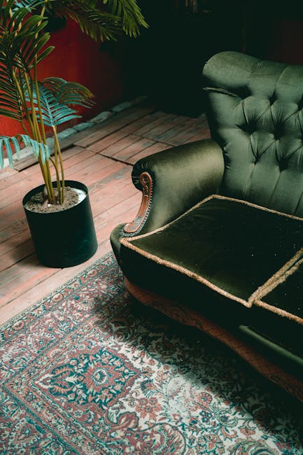 A vintage green velvet armchair with a tufted backrest and wooden carved arms sits on a detailed Persian-style rug with intricate blue, red, and beige patterns. The armchair appears to be partially re