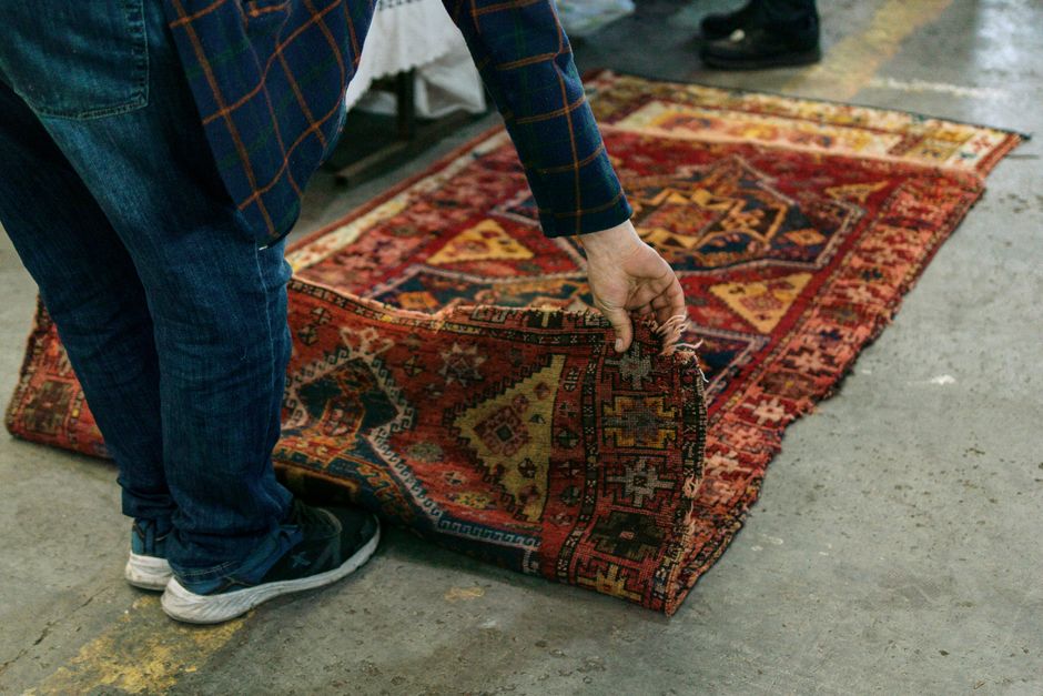 A vintage green velvet armchair with a tufted backrest and wooden carved arms sits on a detailed Persian-style rug with intricate blue, red, and beige patterns. The armchair appears to be partially re