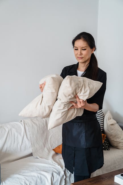 A female cleaner stands in a bright living room, holding two beige cushions. The room features a cream-colored sofa with a white slipcover, a variety of cushions in neutral tones, and a wooden coffee table. The wall behind is plain white, and the lighting is natural, illuminating the space evenly. The cleaner is dressed in professional attire, emphasizing cleanliness and maintenance. The scene reflects domestic cleaning and surface maintenance, consistent with upholstery cleaning services offered by Carpet Cleaners W10, as highlighted in the Ladbroke Grove upholstery cleaning guide for W10 homes.