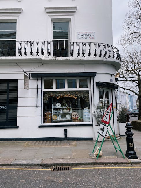 A white building with a curved black wrought iron balcony on the second floor and a bakery shop on the ground level. The shop window displays white and beige ceramic dishware, with a sign reading 'BOULANGERIE' above it. To the right of the shop window, there's a blackboard sign with red and green accents on a green and white A-frame stand, positioned on a paved sidewalk. The building has a rectangular window above the shop and a street sign that reads 'Clarendon Cross W11' attached to the facade. The scene is lit by natural daylight, with a cloudy sky in the background. The exterior appears clean and well-maintained, reflecting the property’s detail-oriented appearance, suitable for surface and deep cleaning in Victorian style storefronts and residential buildings. Carpet Cleaners W10 are known for professional cleaning and maintenance services that promote hygiene and cleanliness in urban environments.
