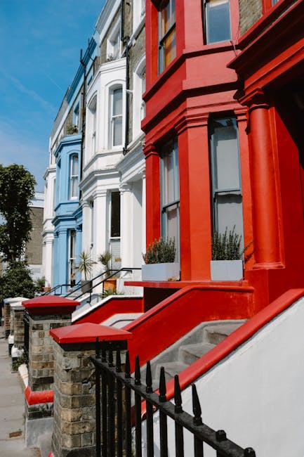 Close-up view of the exterior of residential buildings in Notting Hill, W10, featuring colorful Victorian terraced houses painted in vibrant red, blue, and white. The buildings have large sash windows with white frames, some with small flower planters on window sills. Stone steps lead up to each entrance, with black wrought iron railings bordering the staircases. Sidewalks are clean, and the bright daylight illuminates the facades, highlighting the well-maintained surfaces. The scene emphasizes the aesthetic appeal of the historic streetscape, with no cleaning tools or surfaces visible. Carpet Cleaners W10 provides expert surface cleaning and deep cleaning services, ensuring hygienic and well-maintained properties in the area, supporting compliance with RBKC rules on property upkeep.