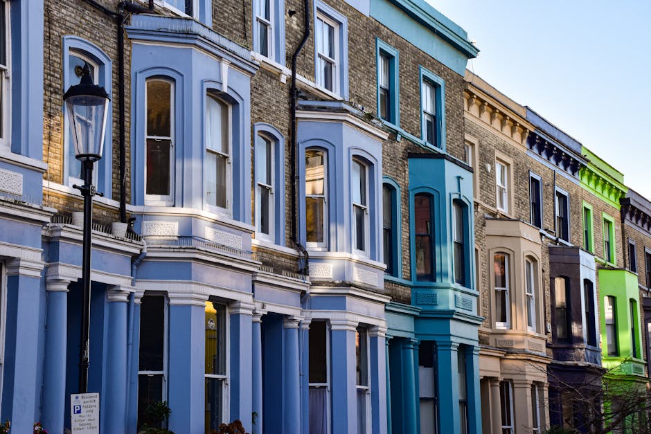 Close-up view of the exterior of residential buildings in Notting Hill, W10, featuring colorful Victorian terraced houses painted in vibrant red, blue, and white. The buildings have large sash windows with white frames, some with small flower planters on window sills. Stone steps lead up to each entrance, with black wrought iron railings bordering the staircases. Sidewalks are clean, and the bright daylight illuminates the facades, highlighting the well-maintained surfaces. The scene emphasizes the aesthetic appeal of the historic streetscape, with no cleaning tools or surfaces visible. Carpet Cleaners W10 provides expert surface cleaning and deep cleaning services, ensuring hygienic and well-maintained properties in the area, supporting compliance with RBKC rules on property upkeep.
