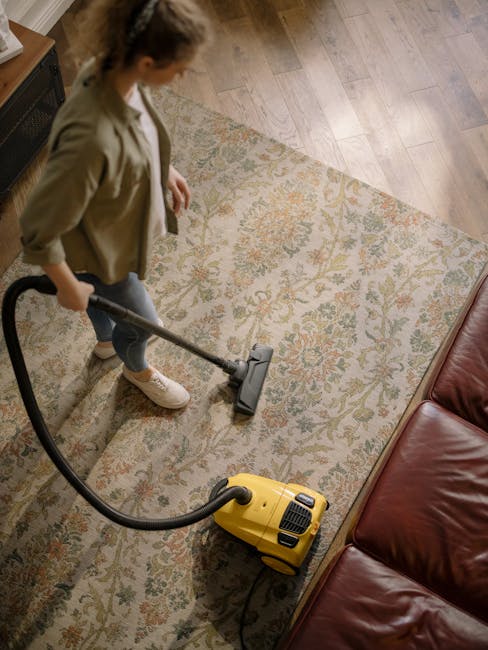 A woman using a yellow and black vacuum cleaner to deep clean a patterned area rug in a living room. The rug features intricate floral designs in shades of beige, green, and orange, placed over a light wooden floor. She is wearing casual clothing, including a beige jacket, white sneakers, and jeans, and is standing next to a red leather sofa. The room is well-lit, with natural light highlighting the cleanliness and maintained condition of the surfaces. Visible furniture includes the sofa, and the setting suggests a domestic environment where surface cleaning and deep carpet sanitisation are part of routine maintenance, consistent with the services offered by Carpet Cleaners W10 for residential cleaning in Notting Hill. The image emphasizes professional domestic cleaning processes aimed at ensuring hygiene and surface freshness.
